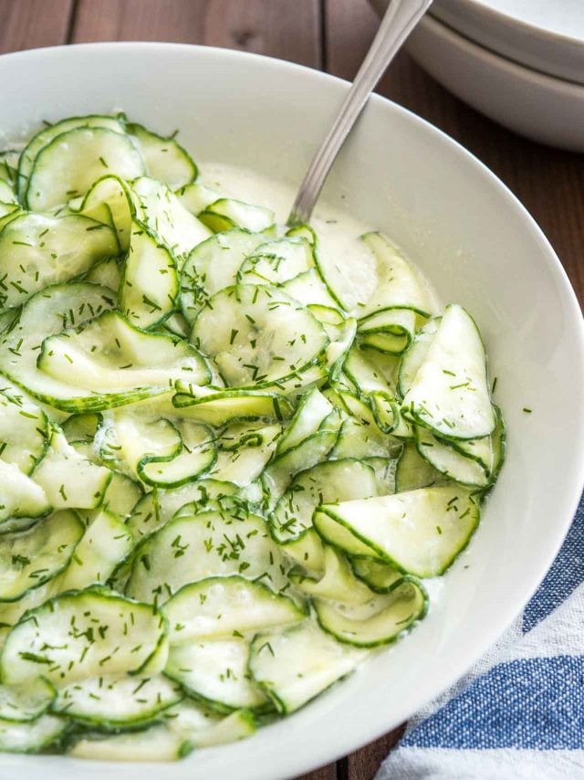 Close-up of German cucumber salad in a white salad bowl with a spoon in it on a white and blue dish towel