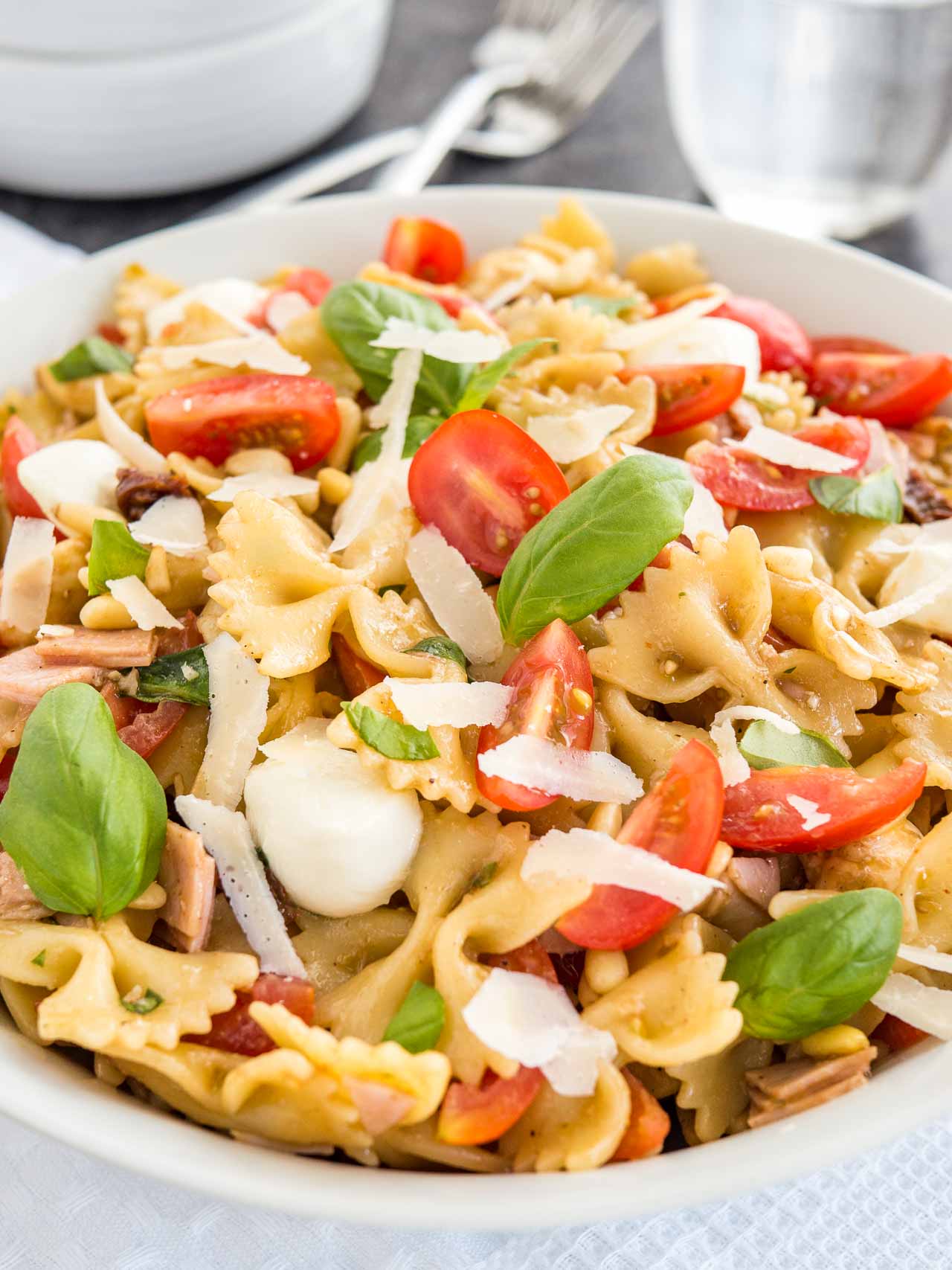 Close-up of a white bowl with pasta salad with Italian dressing, with tomato, mozzarella, basil leaves and shavings of Parmiggiano. The bowl is sitting on a white dishtowel with forks, a glass of water and white bowls.