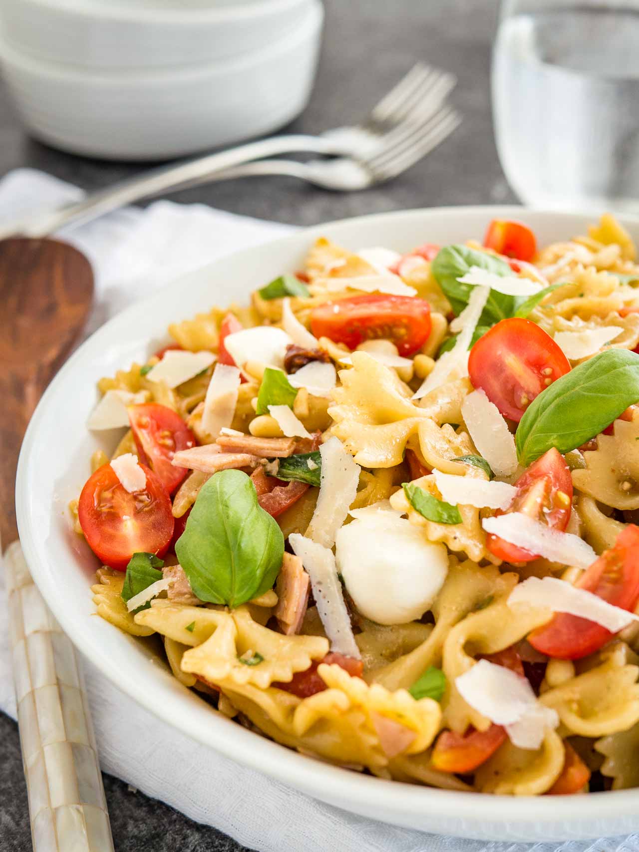 Close-up of a white bowl with pasta salad with Italian dressing, with tomato, mozzarella, basil leaves and shavings of Parmiggiano. The bowl is sitting on a white dishtowel with forks, a glass of water and salad tongs in the background.