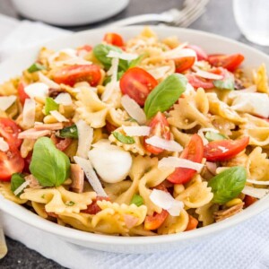 A white bowl with pasta salad with italian dressing, with tomato, mozzarella, basil leaves and shavings of Parmiggiano. The bowl is sitting on a white dishtowel with forks, a glass of water and salad tongs in the background.