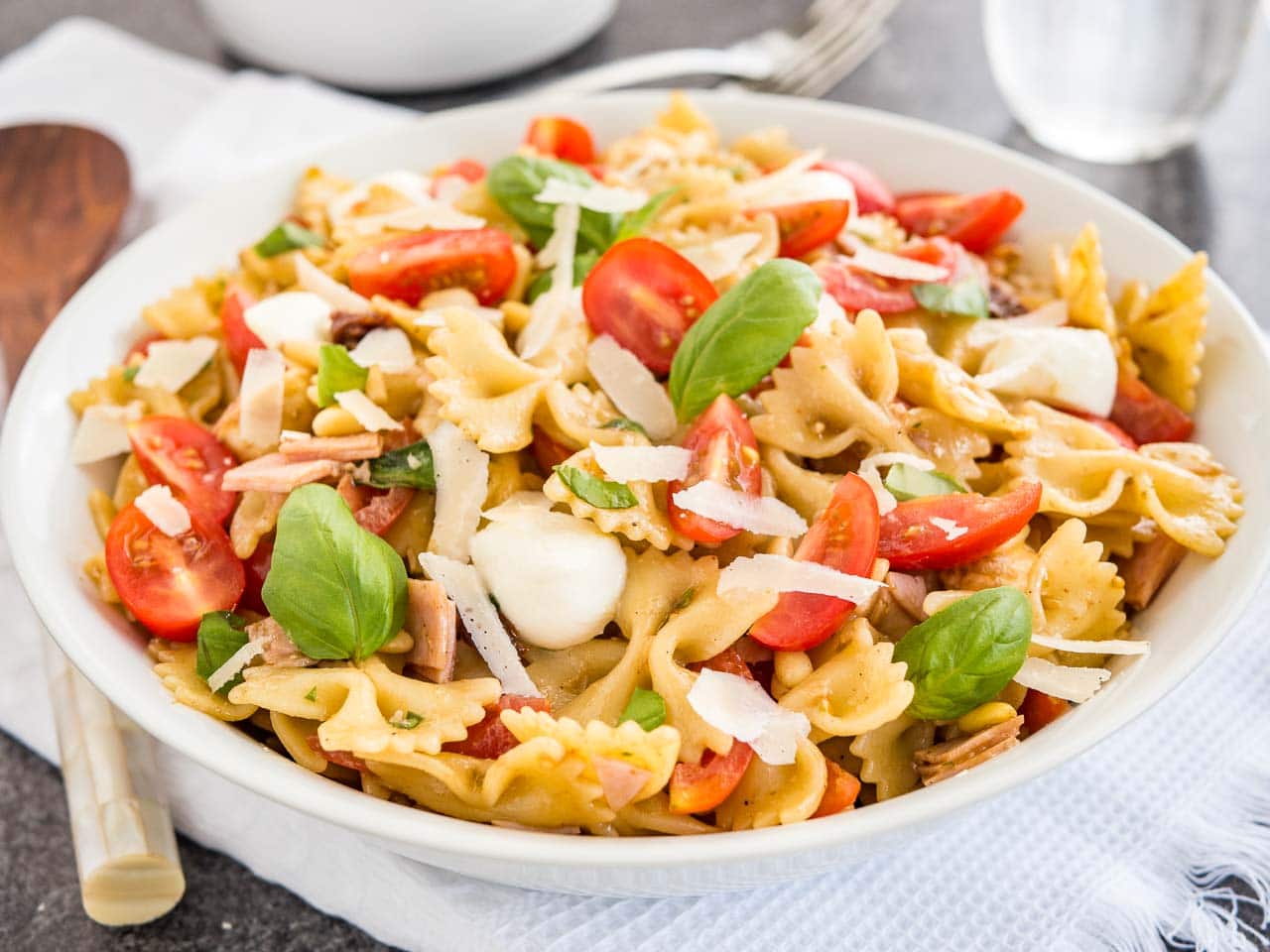 A white bowl with pasta salad with italian dressing, with tomato, mozzarella, basil leaves and shavings of Parmiggiano. The bowl is sitting on a white dishtowel with forks, a glass of water and salad tongs in the background.