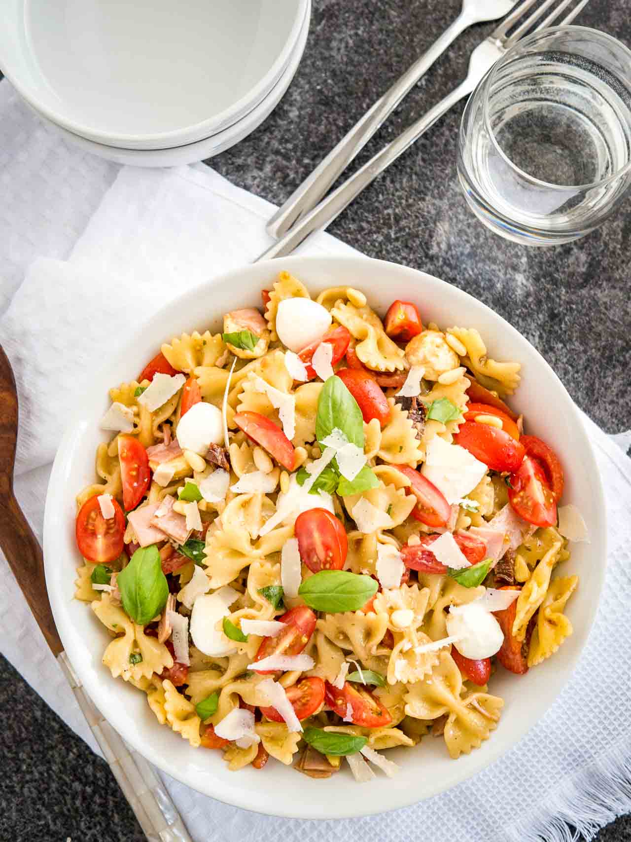 Top-down shot of a white bowl with pasta salad with italian dressing, with tomato, mozzarella, basil leaves and shavings of Parmiggiano. The bowl is sitting on a white dishtowel with forks, a glass of water and salad tongs next to it.