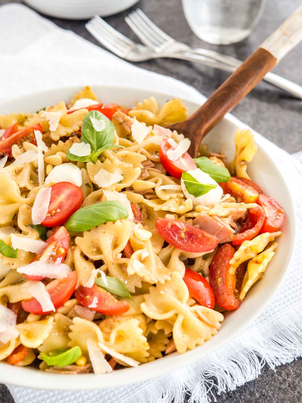 A white bowl with pasta salad with italian dressing, with tomato, mozzarella, basil leaves and shavings of Parmiggiano with a salad tong in it. The bowl is sitting on a white dishtowel with forks next to it.