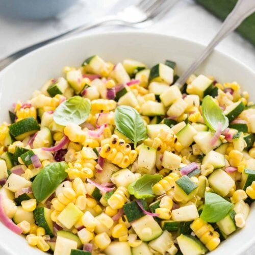 Roasted corn zucchini salad with onions in a white bowl, garnished with basil, with a spoon in it