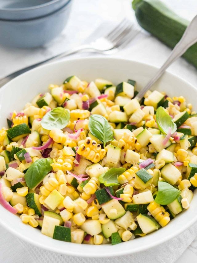 Roasted corn zucchini salad with onions in a white bowl, garnished with basil, with a spoon in it