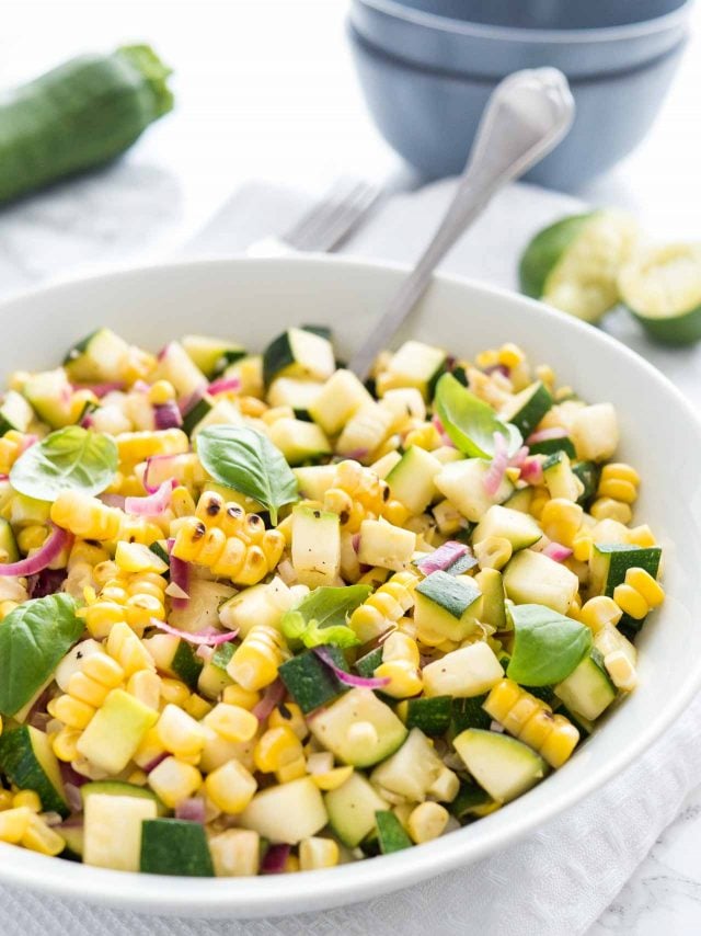 Roasted corn zucchini salad with onions in a white bowl, garnished with basil, with a spoon in it on a white table with a fork, zucchini and squeezed limes in the background