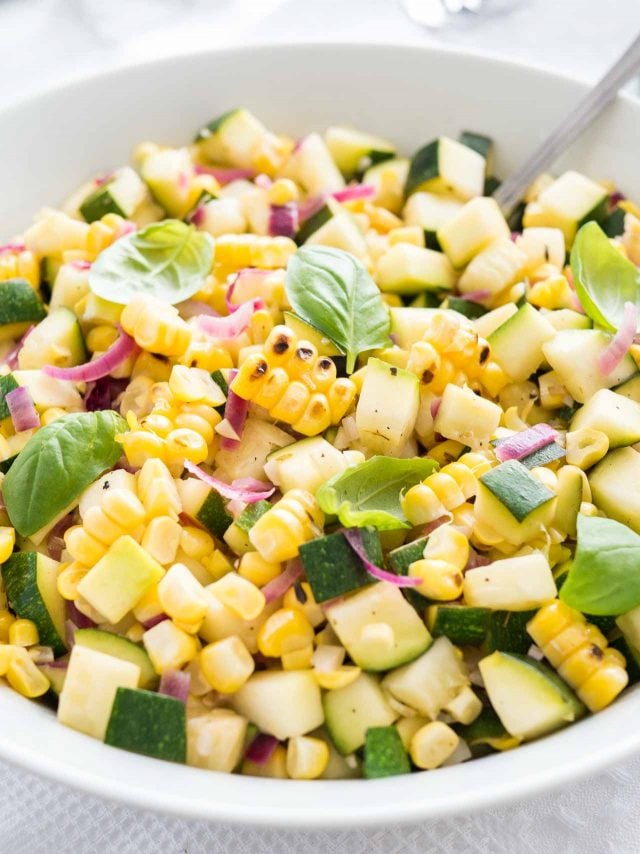 close-up of roasted corn zucchini salad with onions in a white bowl, garnished with basil, with a spoon in it