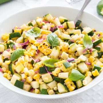 Roasted corn zucchini salad with onions in a white bowl, garnished with basil, with a spoon in it