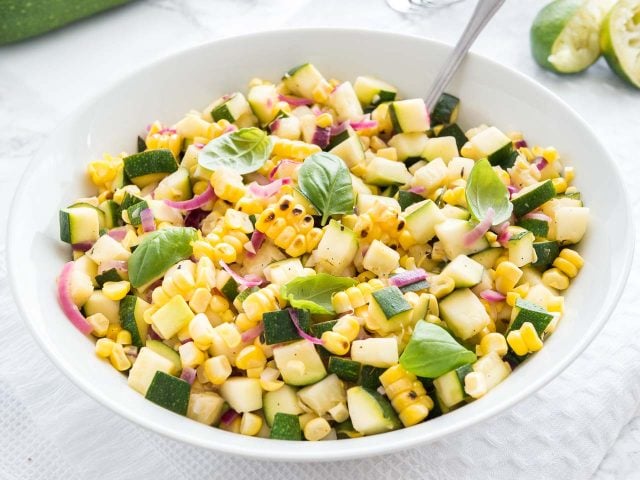 Roasted corn zucchini salad with onions in a white bowl, garnished with basil, with a spoon in it