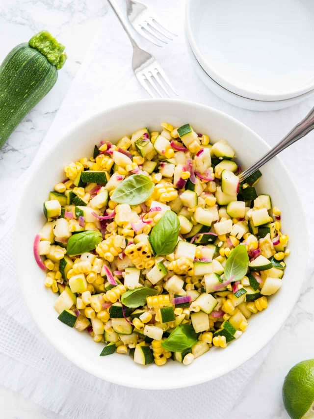 top-down shot of roasted corn zucchini salad with onions in a white bowl, garnished with basil, with a spoon in it on a white table with a fork and a zucchini next to it