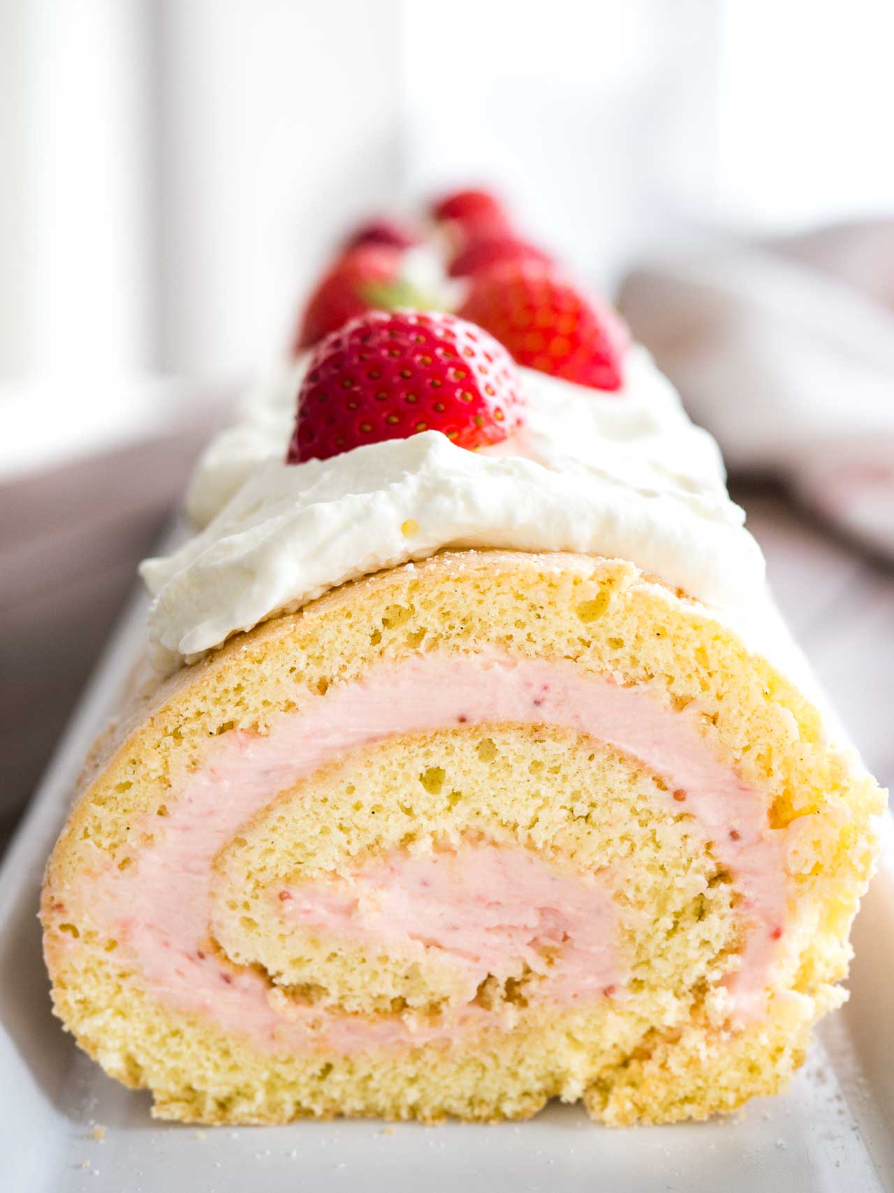 Close-up of a strawberry swiss cake roll topped with whipped cream and strawberries on a rectangular serving platter.