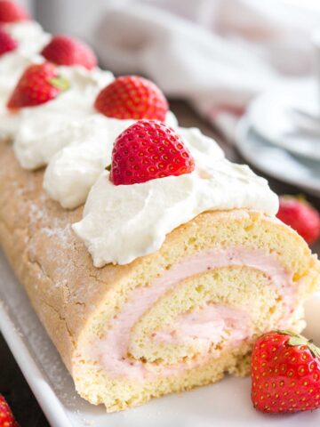 Close-up of a strawberry swiss cake roll topped with whipped cream and strawberries on a rectangular serving platter.