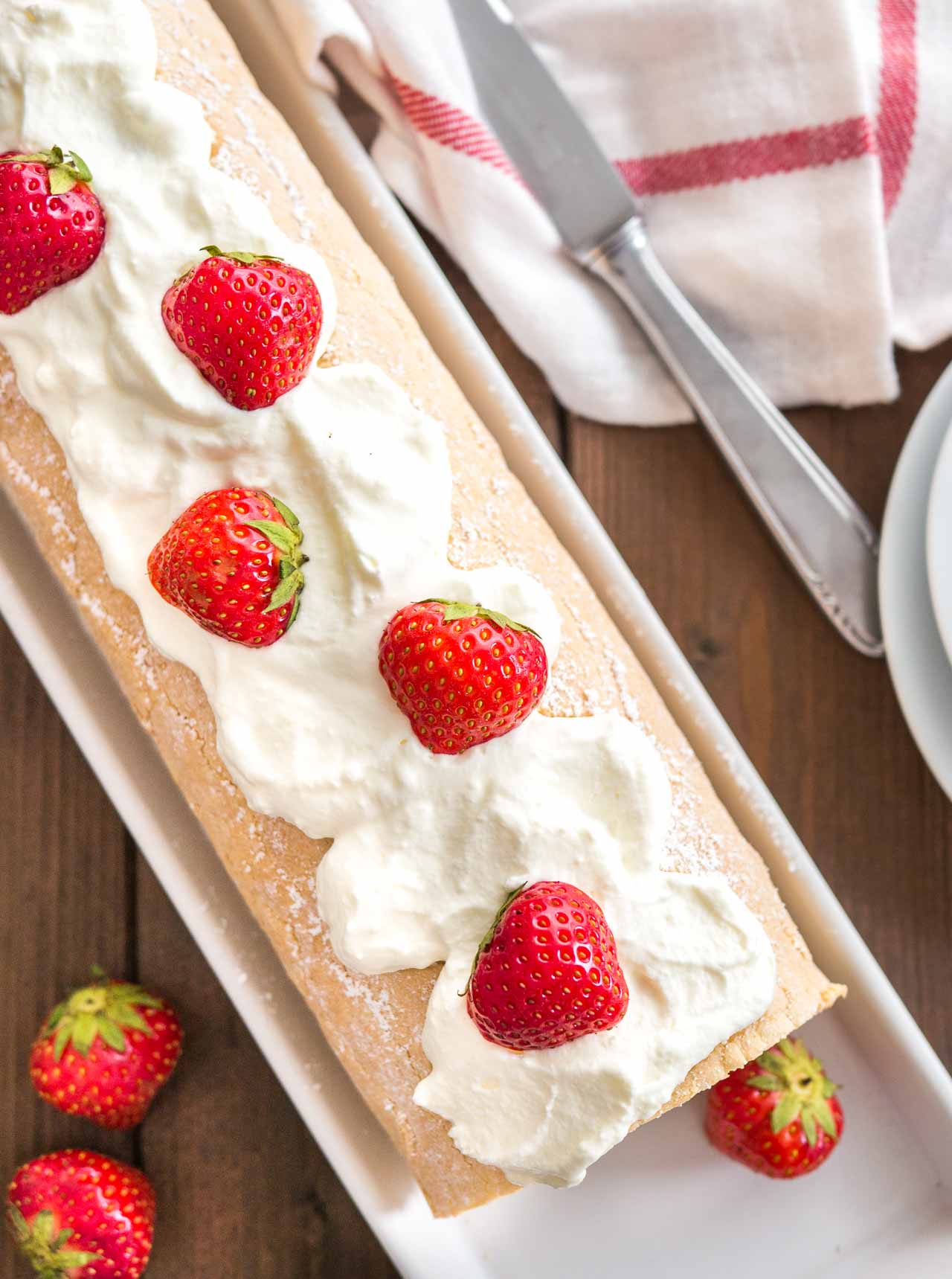 Top-down shot of a strawberry swiss cake roll topped with whipped cream and strawberries on a rectangular serving platter. There are plates, a knife and a white and red dishtowel next to it.