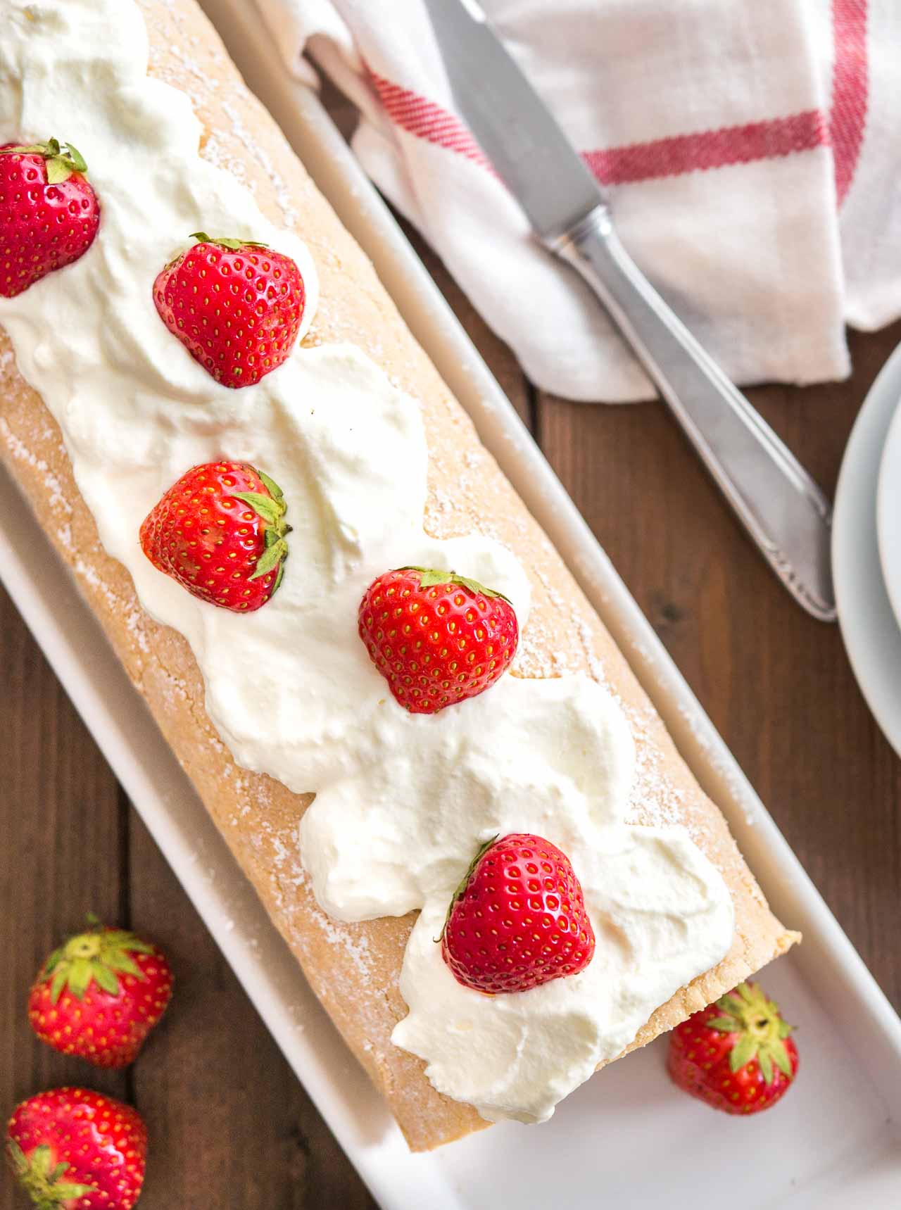 Top-down shot of a strawberry roll cake topped with whipped cream and strawberries on a rectangular serving platter. There are plates, a knife and a white and red dishtowel next to it.