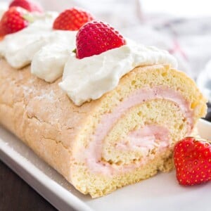 A strawberry swiss cake roll topped with whipped cream and strawberries on a rectangular serving platter.