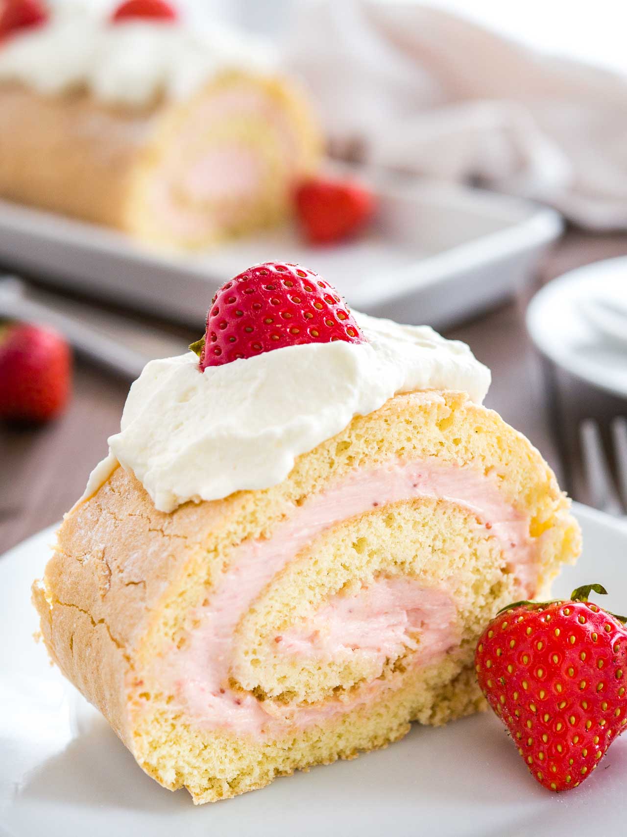 A slice of strawberry swiss roll cake on a white plate topped with whipped cream and strawberries. The rest of the roll is in the background.