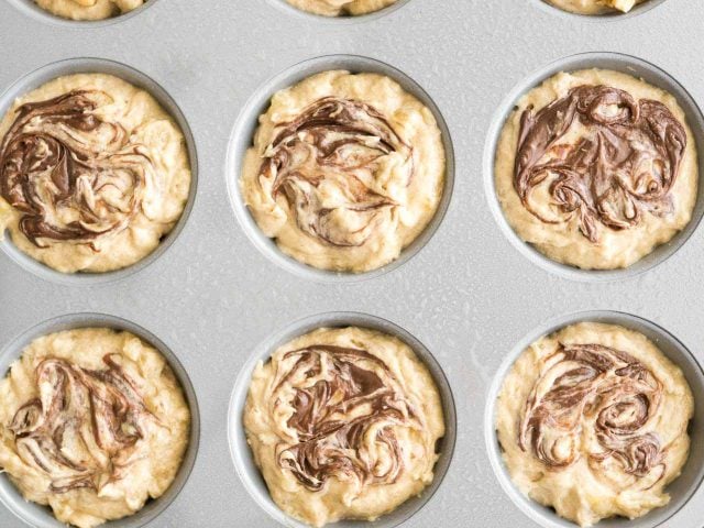 Top-down shot of a grey muffin pan with banana nutella muffin batter in it before baking.