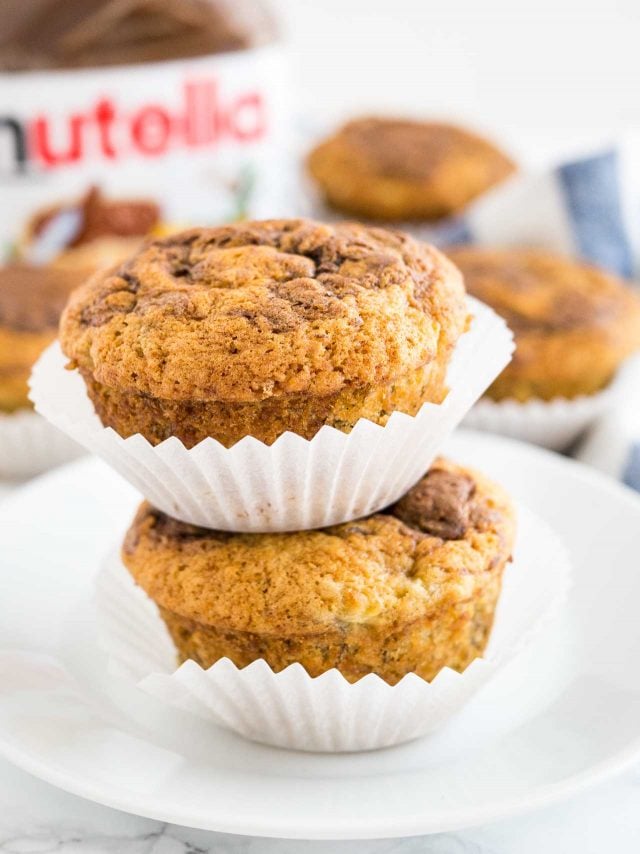 Close-up of two banana nutella muffins stacked on top of each other on a white plate, with more muffins and a jar of nutella in the background.