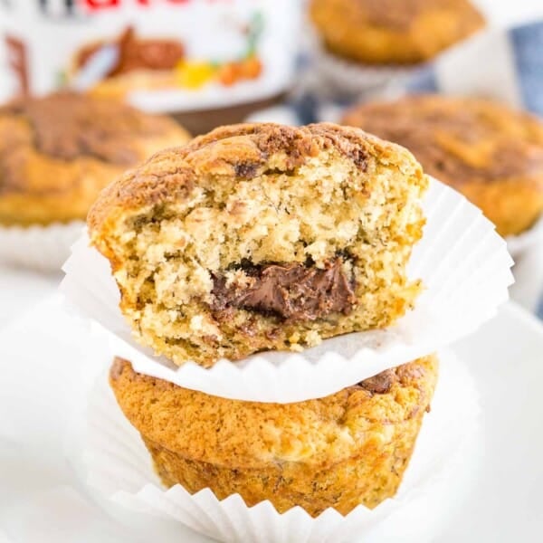 Close-up of two banana nutella muffins stacked on top of each other on a white plate, with more muffins and a jar of nutella in the background. The top muffin has had a big bite taken out of it.