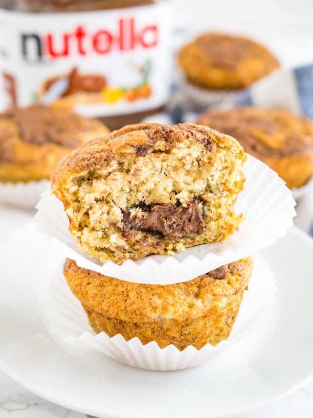 Close-up of two banana nutella muffins stacked on top of each other on a white plate, with more muffins and a jar of nutella in the background. The top muffin has had a big bite taken out of it.