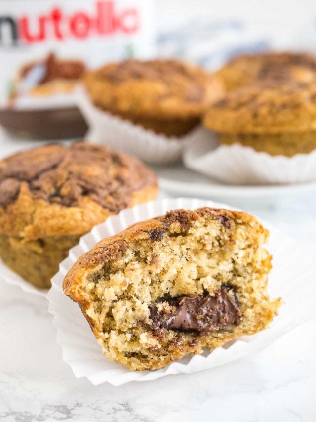 Close-up of a banana nutella muffin sitting in its liner with a big bite taken out of it. There are more muffins and a jar of nutella in the background.