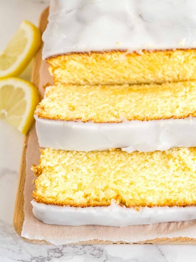 Close-up of a loaf of moist lemon cake on a wooden cutting board, line with parchment paper. Two slices have been cut off and are lying in front of it. Next to it, there are two lemon wedges.