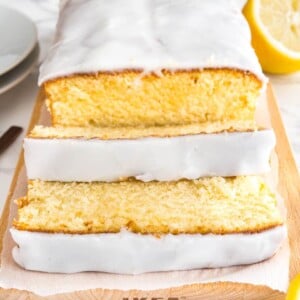 A loaf of moist lemon cake on a wooden cutting board, line with parchment paper. Two slices have been cut off and are lying in front of it. Next to it, there's half a lemon and two white plates.