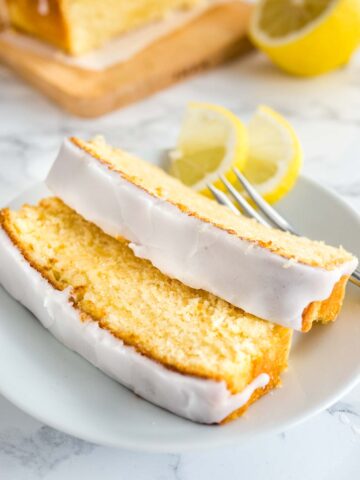 Two slices of moist lemon cake with lemon glazing lying on a white plate with a fork garnished with two lemon wedges. A wooden cutting board with the rest of the cake is in the background.