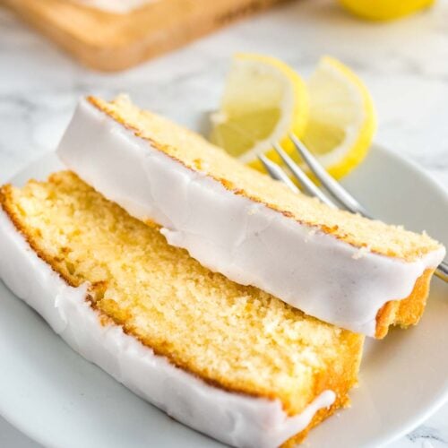 Two slices of moist lemon cake with lemon glazing lying on a white plate with a fork garnished with two lemon wedges. A wooden cutting board with the rest of the cake is in the background.