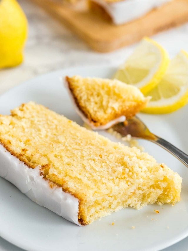 A slice of moist lemon cake with lemon glazing lying on a white plate garnished with two lemon wedges. A fork has taken out a piece of it. There\'s a wooden cutting board and half a lemon in the background.