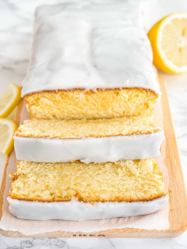 A loaf of moist lemon cake on a wooden cutting board, line with parchment paper. Two slices have been cut off and are lying in front of it. Next to it, there\'s half a lemon and two lemon wedges next to it.
