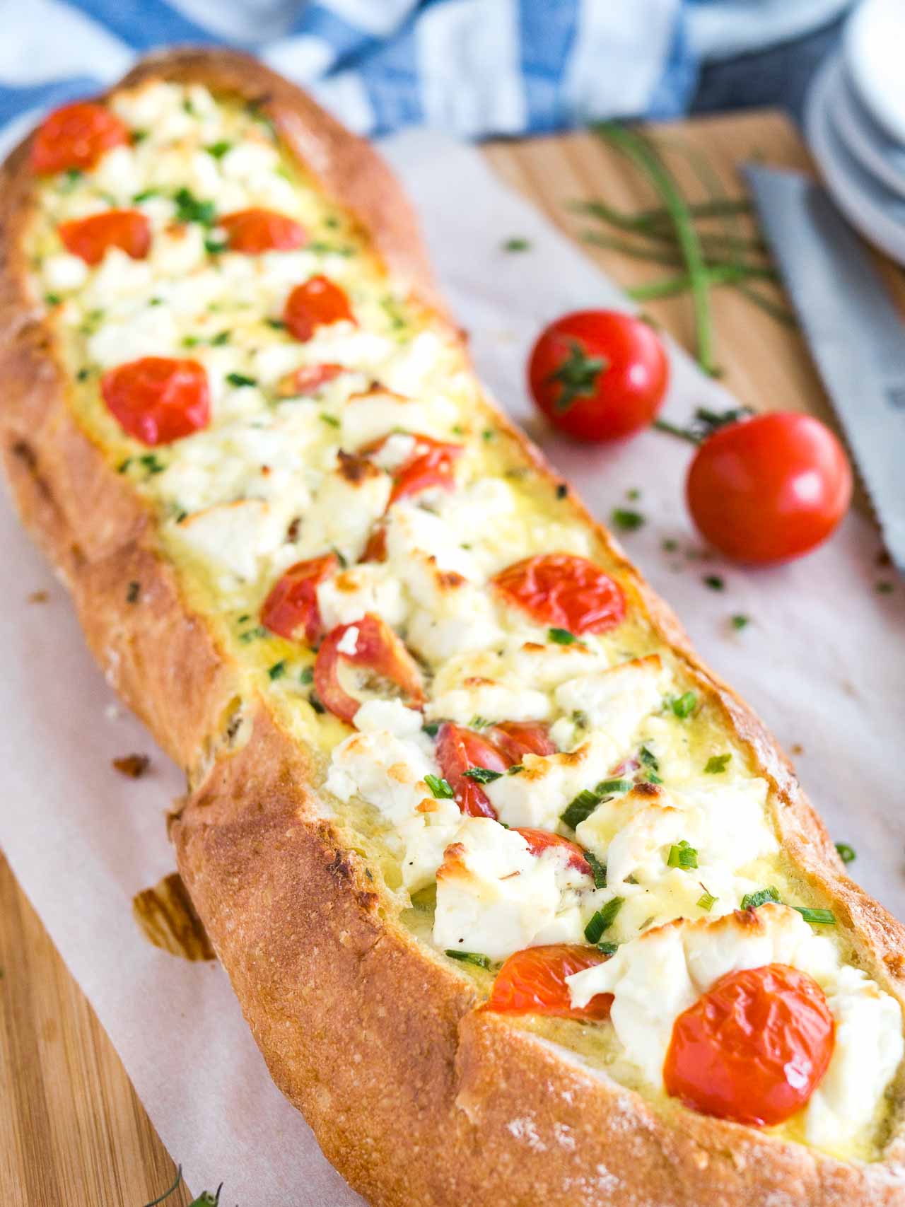 Close-up of tomato feta stuffed french bread garnished with tomatoes and chives on a bamboo cutting board lined with parchment paper. There\'s a bread knife lying next to it.