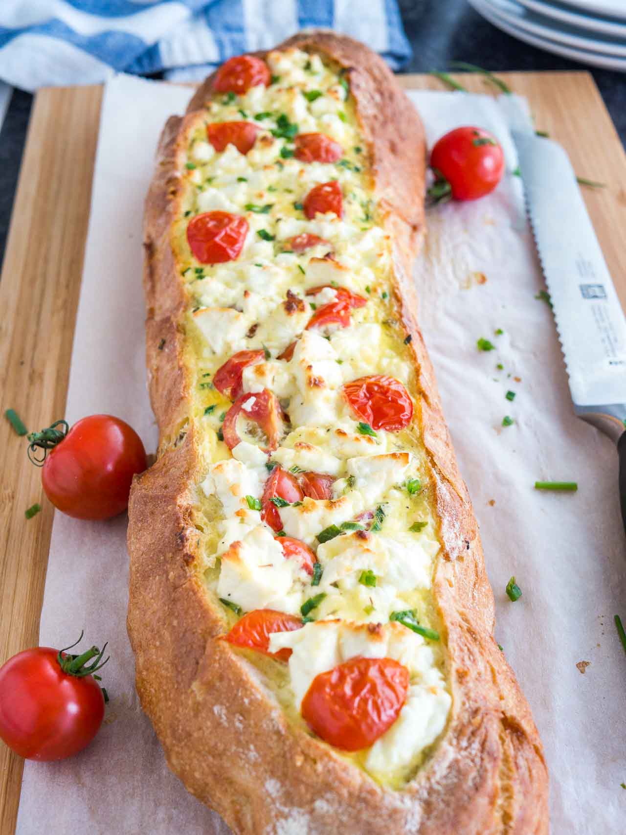 Tomato feta stuffed french bread garnished with tomatoes and chives on a bamboo cutting board lined with parchment paper. There\'s a bread knife lying next to it.