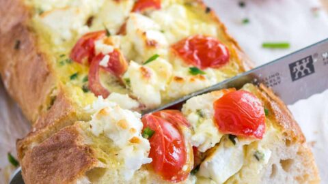 A bread knife cutting into tomato feta stuffed french bread garnished with tomatoes and chives on a bamboo cutting board lined with parchment paper.