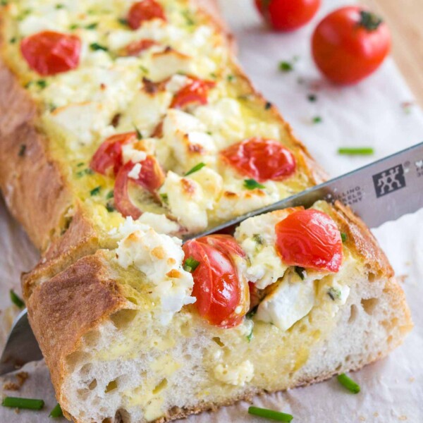 A bread knife cutting into tomato feta stuffed french bread garnished with tomatoes and chives on a bamboo cutting board lined with parchment paper.