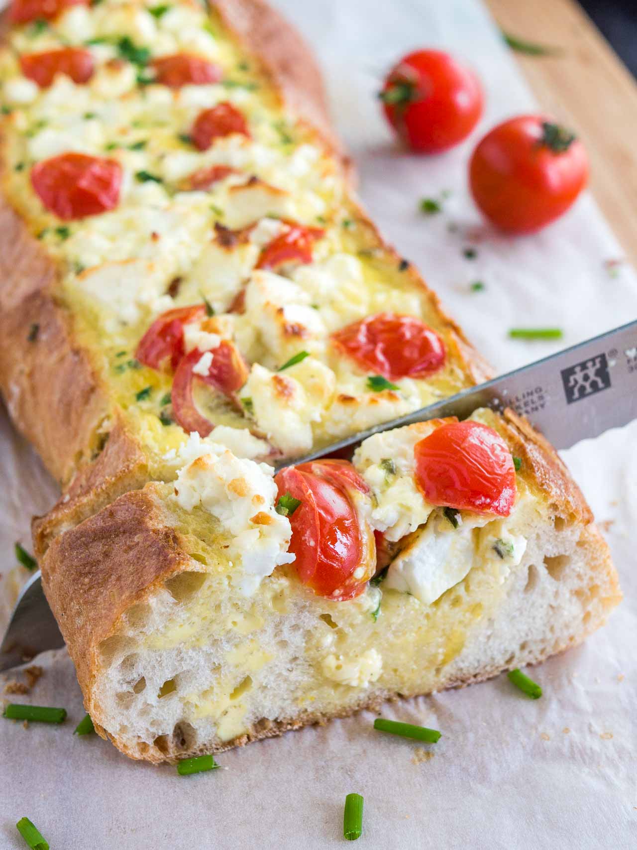 A bread knife cutting into tomato feta stuffed french bread garnished with tomatoes and chives on a bamboo cutting board lined with parchment paper.