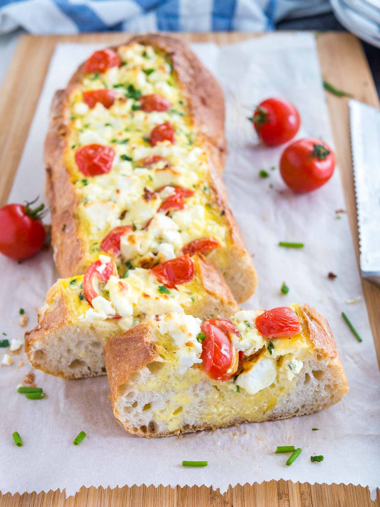 Tomato feta stuffed french bread garnished with tomatoes and chives on a bamboo cutting board lined with parchment paper. Two slices have been cut off and are sitting in front of it and there\'s a bread knife lying next to it.