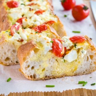 Tomato feta stuffed french bread garnished with tomatoes and chives on a bamboo cutting board lined with parchment paper. A slice has been cut off and is sitting in front of it.