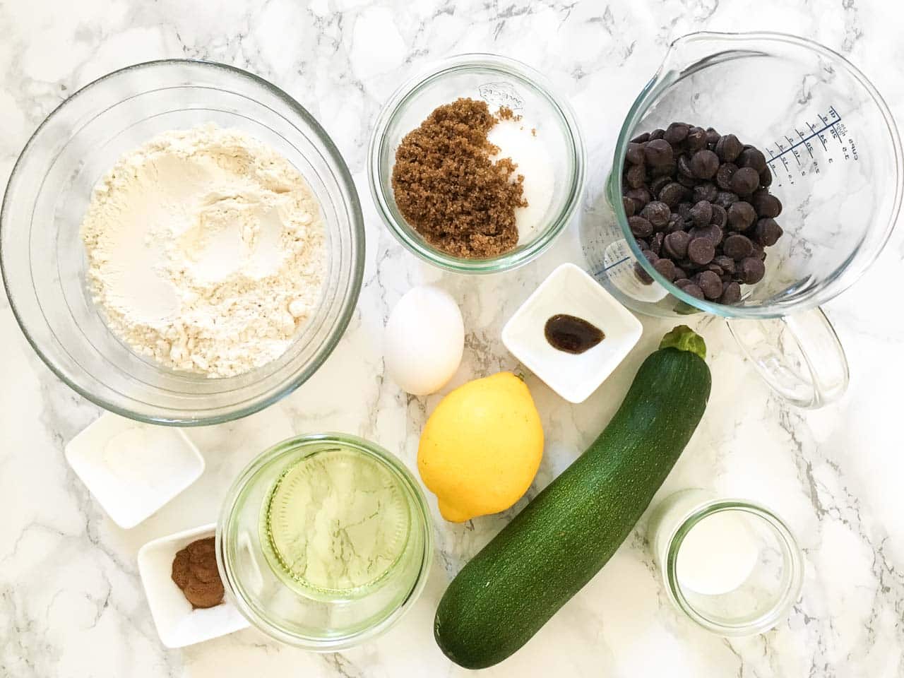 Top-down shot of ingredients in various bowls: Zucchini, flour, egg, white and brown sugar, oil, mil, lemon juice, vanilla extract, baking soda, baking powder, cinnamon, salt, chocolate chips.