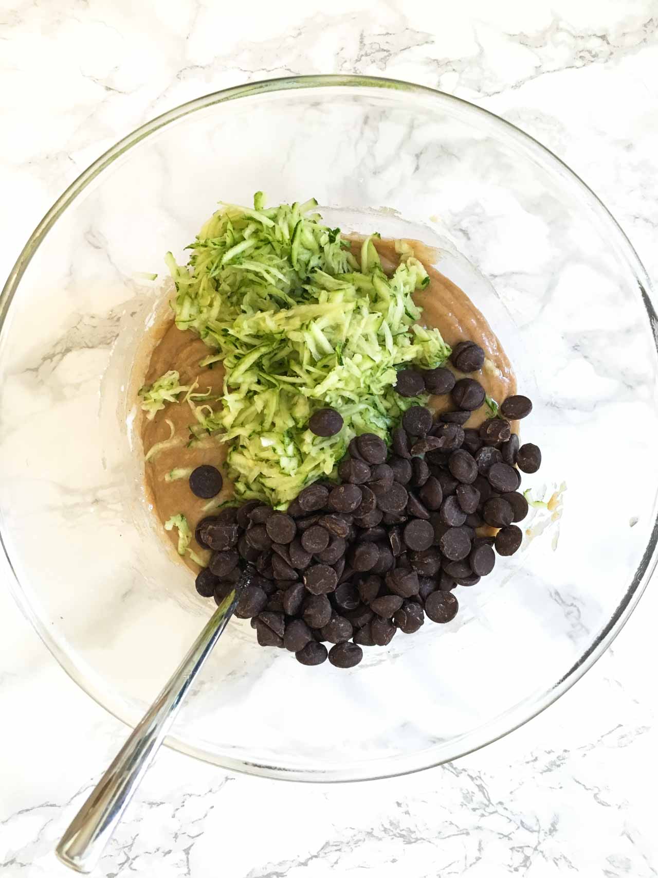 Top-down shot of a glass bowl of muffin batter with shredded zucchini and chocolate chips on top. The bowl has a spoon in it and is sitting on a marble surface.