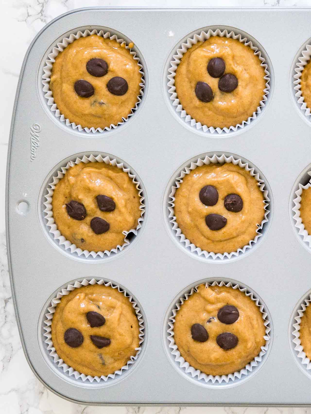 Top-down shot of a grey muffin pan with unbaked pumpkin chocolate chip muffins on a marble surface.
