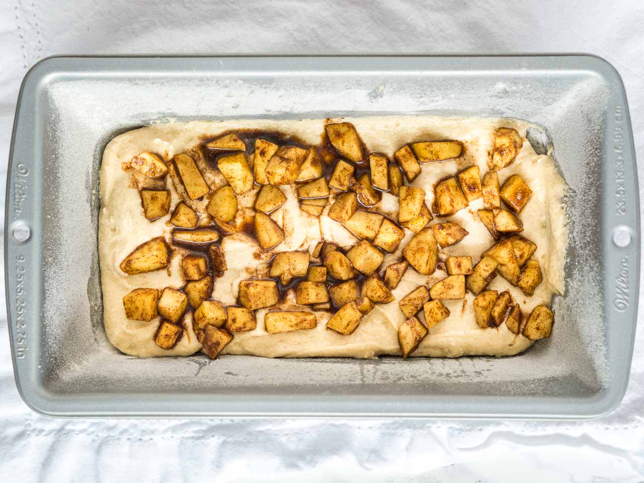 A floured bread pan with apple cinnamon bread batter and apple dice with cinnamon and sugar on top.