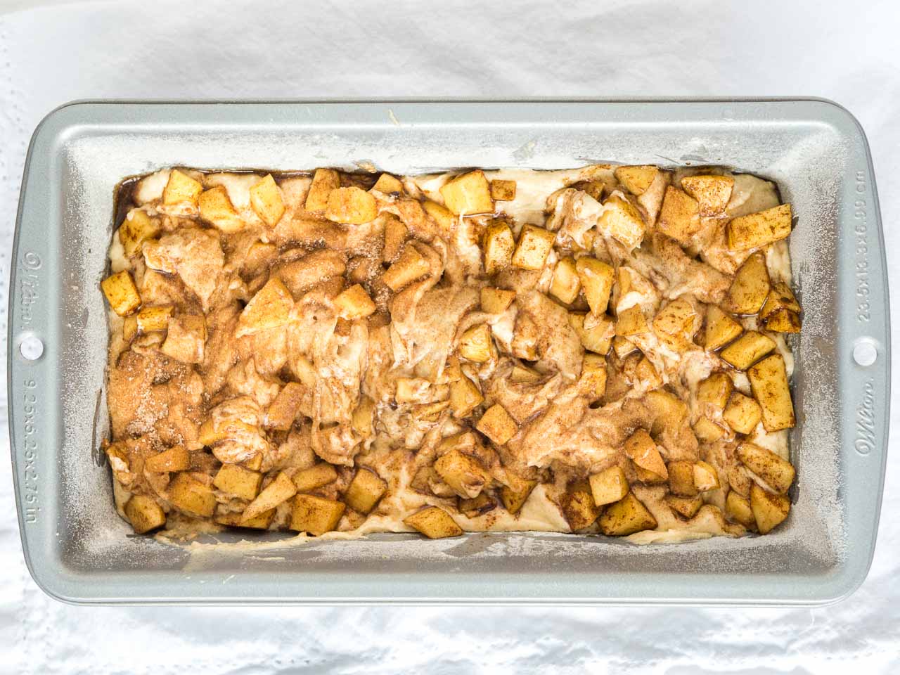 A floured bread pan with apple cinnamon bread batter and apple dice with cinnamon and sugar on mixed together.