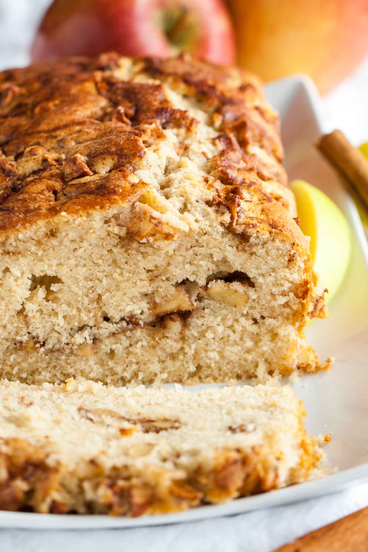 Close-up of a rectangular plate with apple cinnamon bread. A slice has been cut off and is lying in front. Garnished with apple wedges, whole apples and cinnamon sticks.