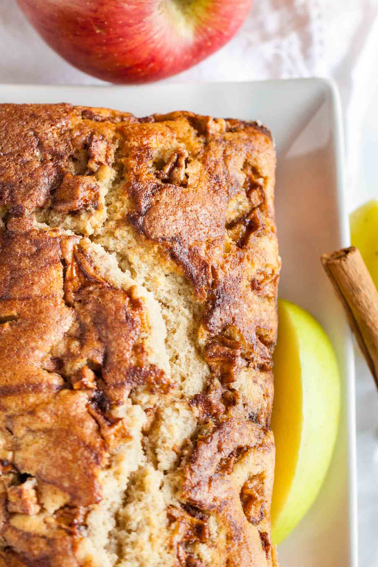 Top-down shot of a rectangular plate with apple cinnamon bread. A slice has been cut off and is lying in front. Garnished with apple wedges, whole apples and cinnamon sticks.