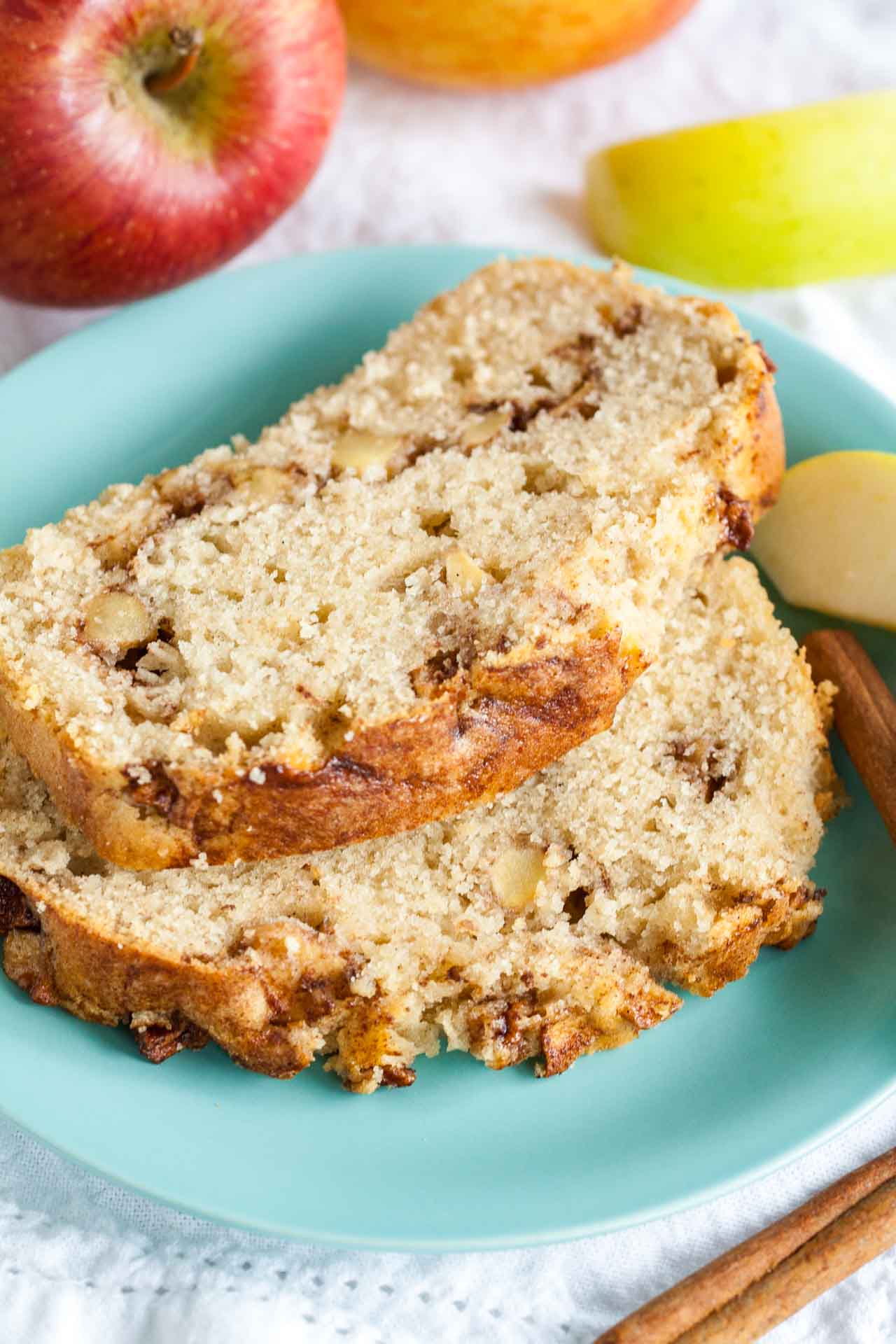 A teal plate with two slices of apple cinnamon bread, garnished with apples and cinnamon sticks on a white dishtowel.