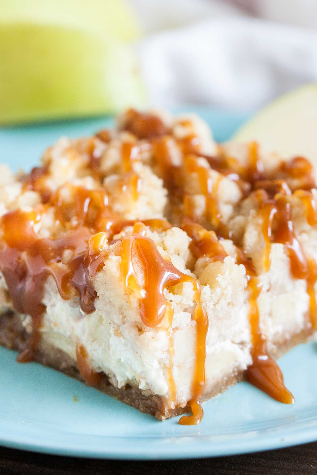 Close-up of a teal plate with a caramel apple cheesecake bar garnished with apples and cinnamon sticks.