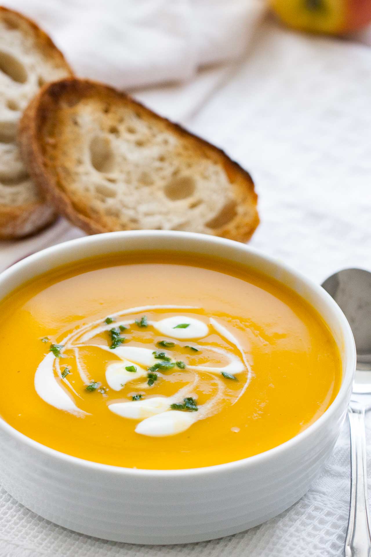 A white bowl with butternut squash soup, with dollops of sour cream and chives. It is sitting on a white dish towel and there\'s a spoon and toasted slices of bread next to it.