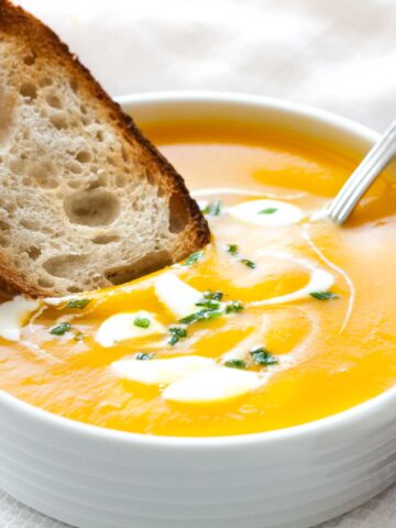 Close-up of a white bowl with butternut squash soup, with dollops of sour cream and chives, a spoon in it and a toasted slice of bread. It is sitting on a white dish towel next to some toasted slices of bread.