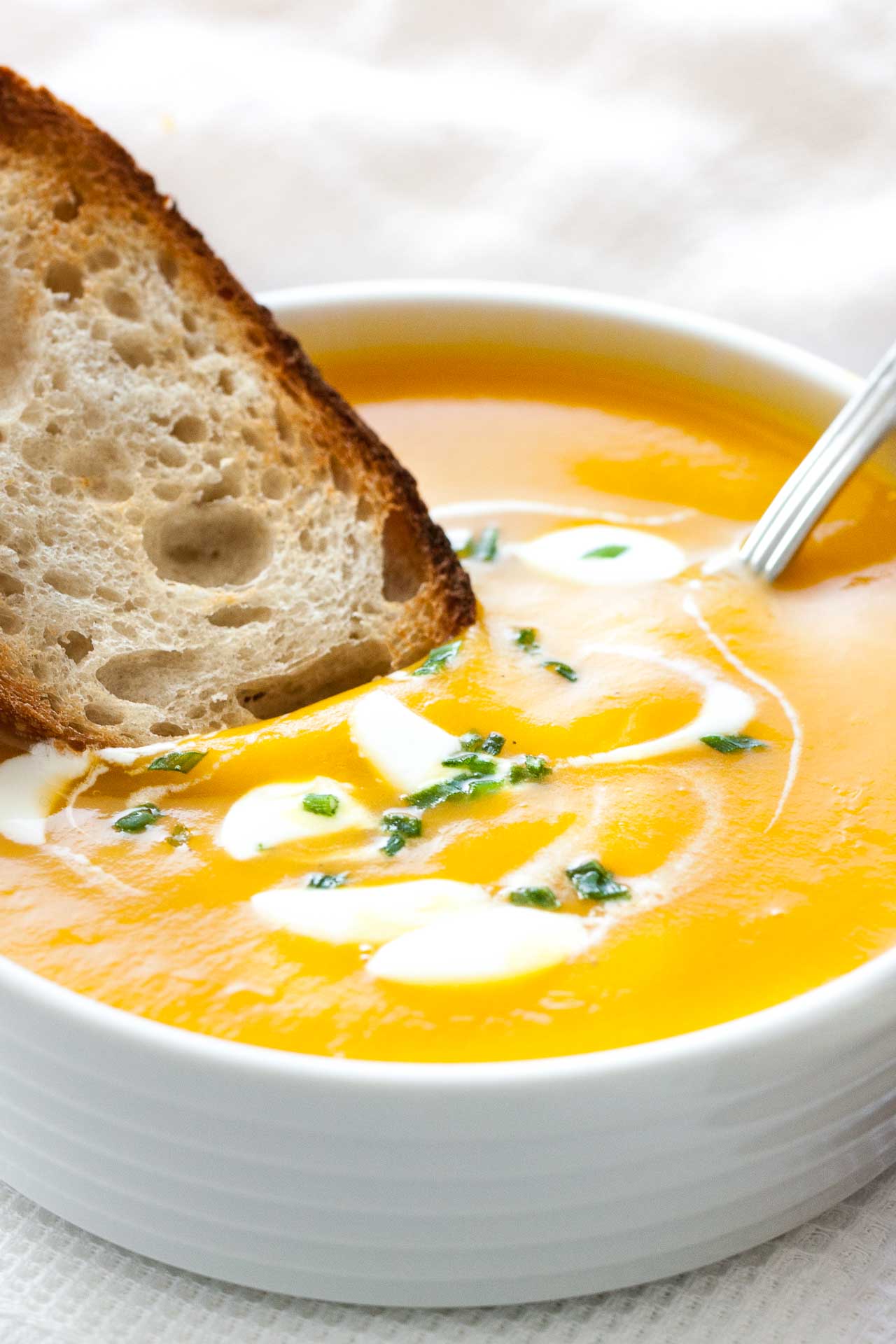 Close-up of a white bowl with butternut squash soup, with dollops of sour cream and chives, a spoon in it and a toasted slice of bread. It is sitting on a white dish towel next to some toasted slices of bread.